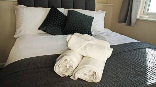 A neatly made bed with white and dark pillows, a folded white towel, and a dark textured bedspread. - Host Stay Clacton Waves (Clacton-on-Sea)