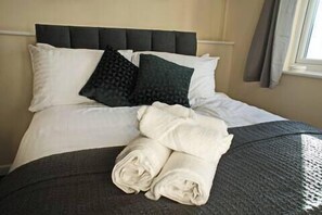 A neatly made bed with white and dark pillows, a folded white towel, and a dark textured bedspread. - Host Stay Clacton Waves (Clacton-on-Sea)