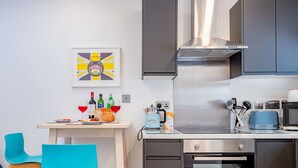 A modern kitchen with a stainless steel range hood, a coffee maker, and a toaster. There is a framed picture on the wall and a table with a loaf of bread and wine glasses. - Host Stay Croydon Road Abode (Caterham)