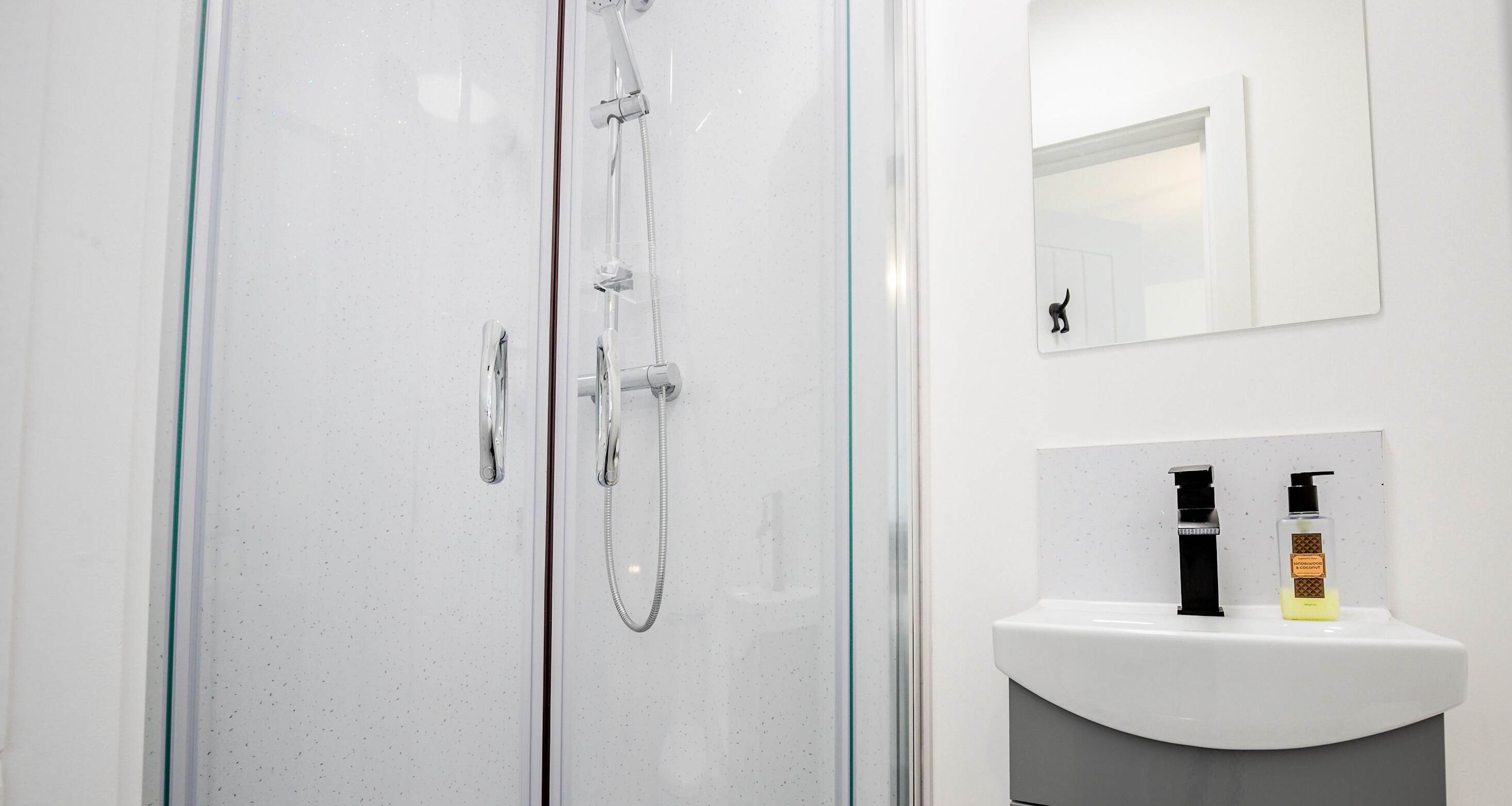 Ein modernes Badezimmer mit einer ein Glaswand trennenden Dusche, einem weißen Waschbecken mit Spiegel darüber und zwei Seifenflaschen auf dem Waschbecken.