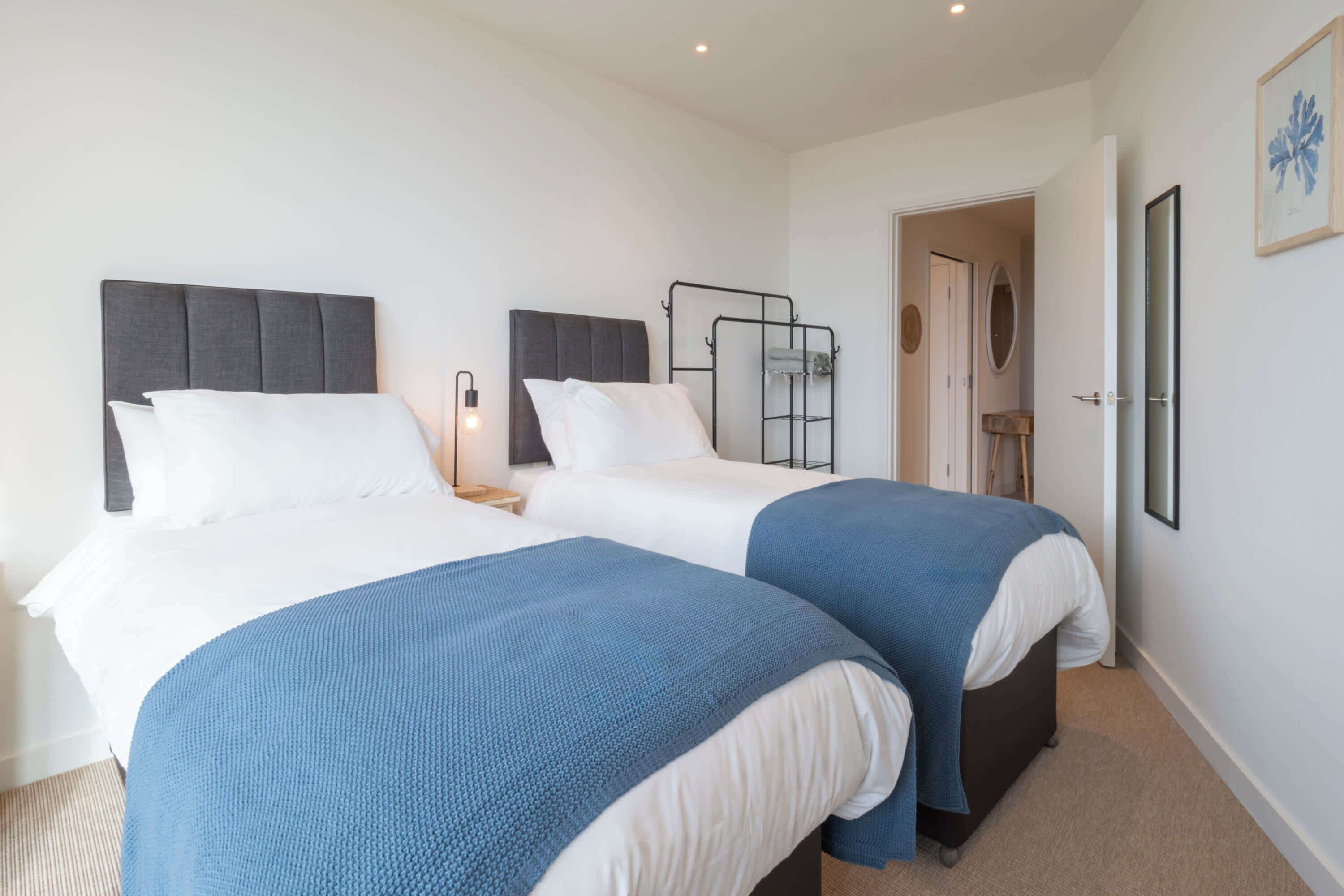 A hotel room with two beds, each with a grey headboard and white bedding. There is a bedside table with a lamp, a door leading to another room, and a framed picture on the wall.