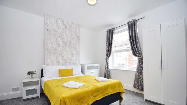 A bedroom with a bed, a nightstand, a wardrobe, a window with curtains, and a wall with a floral pattern. - Host Stay Church Drive (Mansfield)