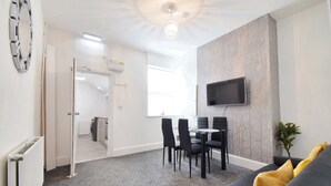 A modern living room with a grey carpet, a black sofa with yellow cushions, a dining area with a table and chairs, a flat-screen TV mounted on the wall, and a radiator on the left. - Host Stay Church Drive (Mansfield)