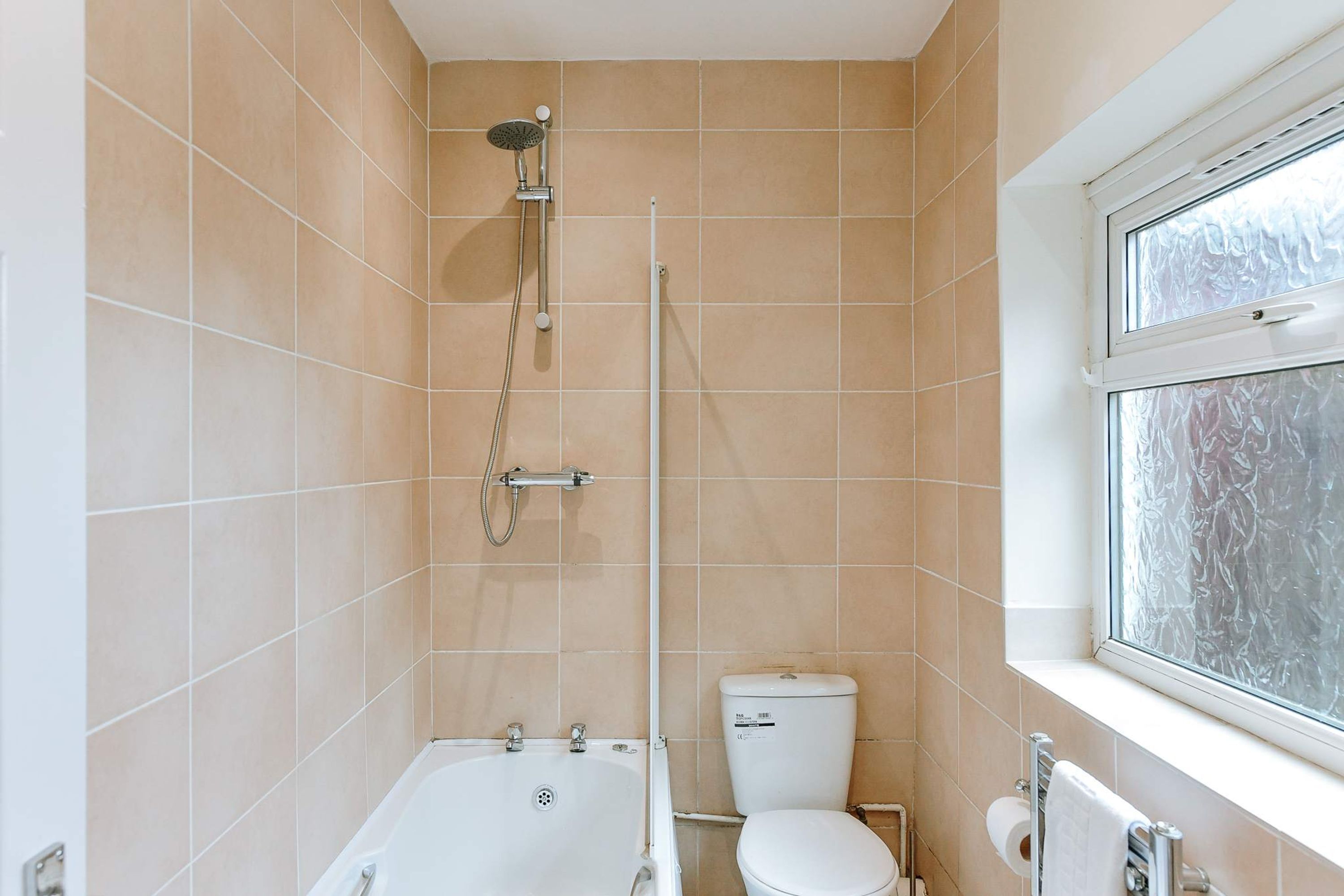 A bathroom with beige tiles, a bathtub, a toilet, and a window with frosted glass.