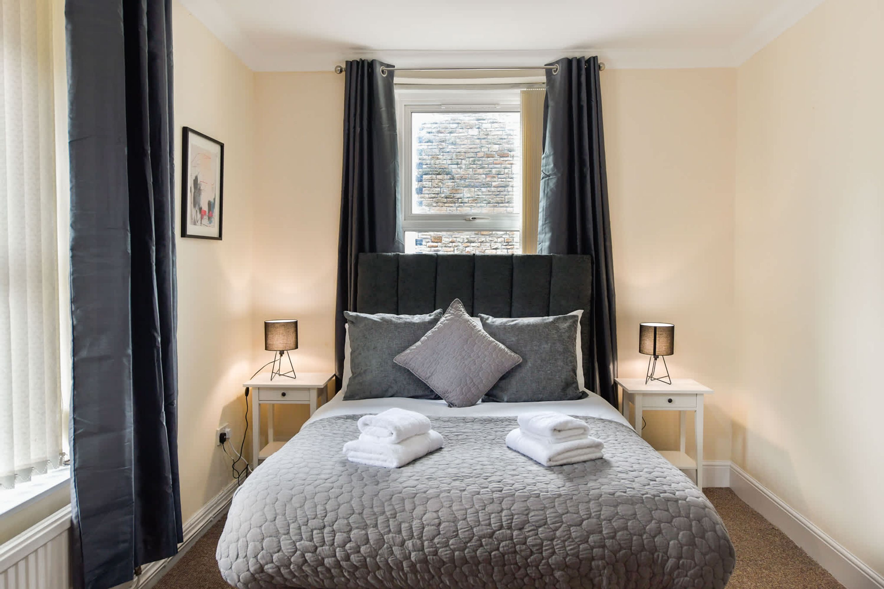 A neatly arranged bedroom with a large bed, two bedside lamps, a window with curtains, and a framed picture on the wall.