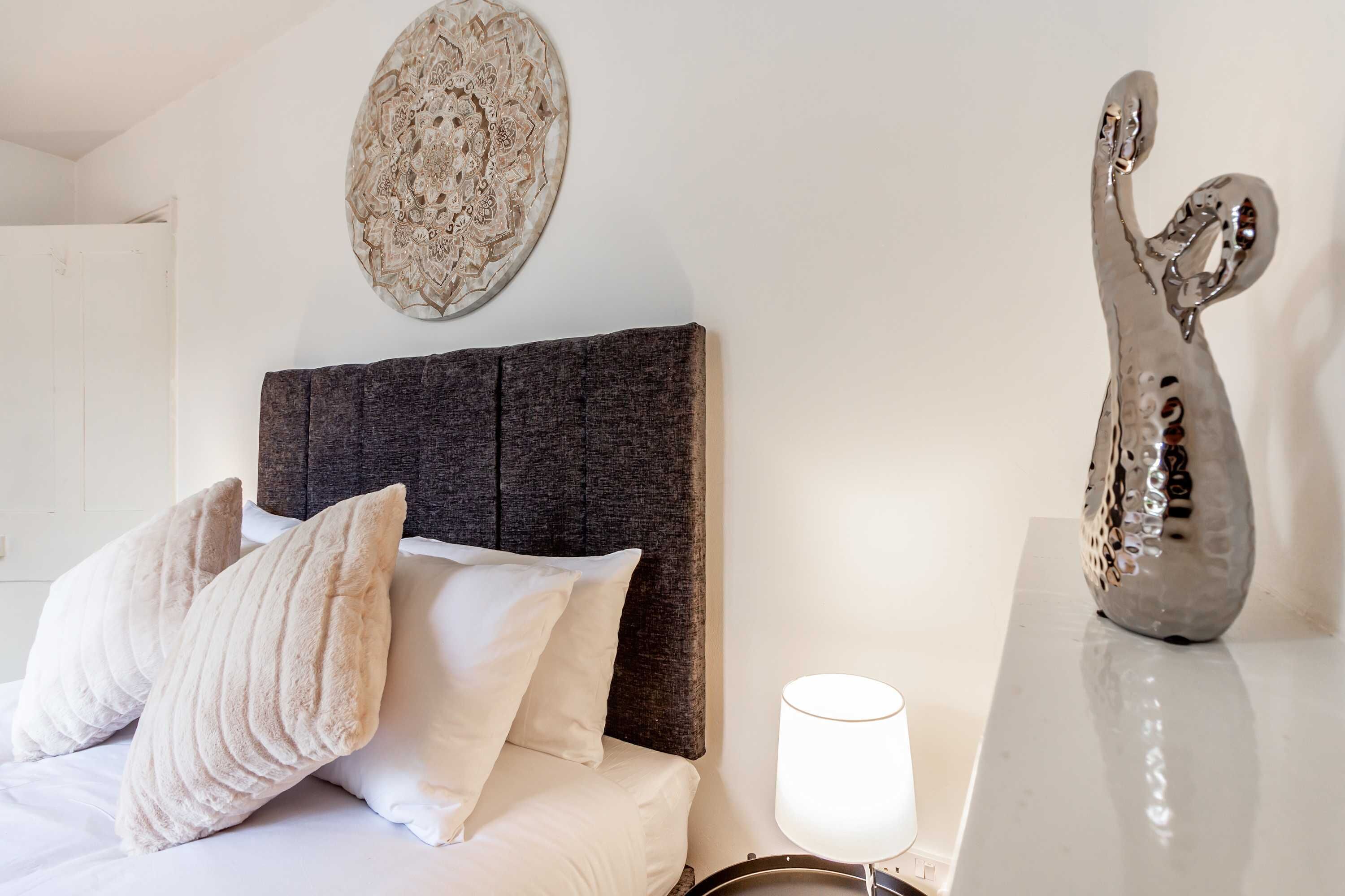 A neatly made bed with white linens and two beige pillows, a dark headboard, a decorative wall piece, and a reflective sculpture on a white table.