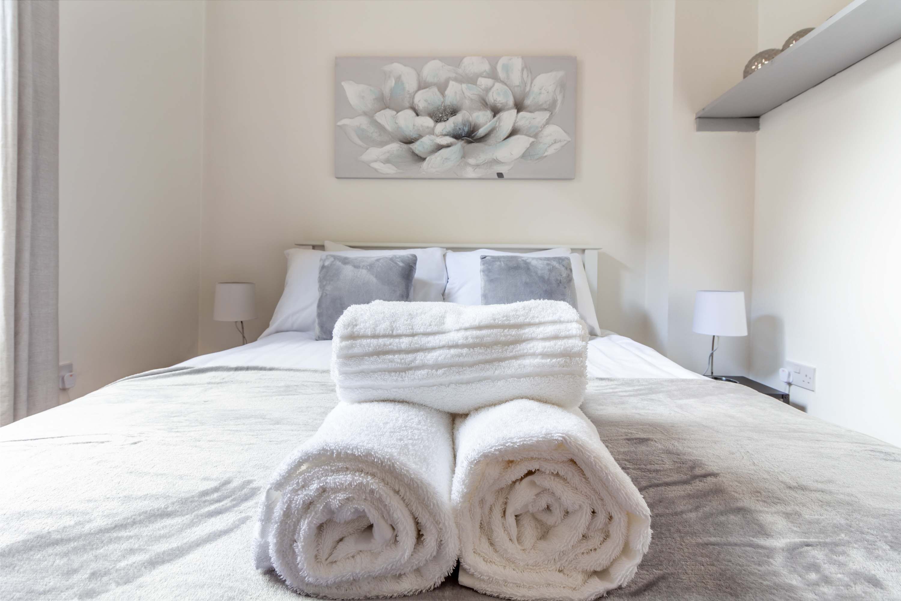 Una cama bien hecha con toallas blancas enrolladas, un cuadro de flores en la pared y dos lámparas de noche.