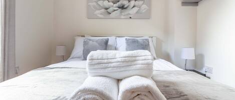 Una cama bien hecha con toallas blancas enrolladas, un cuadro de flores en la pared y dos lámparas de noche.
