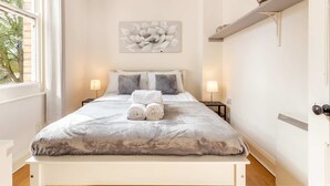A neatly made bed with a grey blanket and two rolled towels, a wall-mounted flower painting, and a shelf with a decorative item. - Host Stay Club Chambers (Malvern)