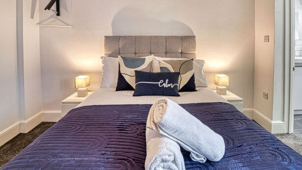 Un lit bien fait, avec une serviette pliée, deux lampes de chevet et une tête de lit agrémentée d’un coussin.