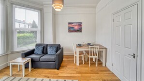 Une pièce avec un canapé gris, une petite table, une table à manger avec des chaises, un tableau au mur et une porte blanche.