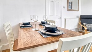 Une table à manger est dressée avec des assiettes, des bols et des verres à vin, entourée de chaises blanches dans une pièce dotée d’une porte blanche et d’un miroir au mur.