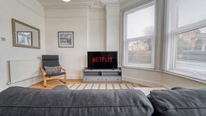 Un salon avec un canapé gris, une chaise en bois, un téléviseur posé sur un meuble et une fenêtre avec des stores.