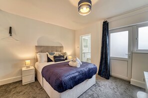 Ein Hotelzimmer mit Bett, Nachttisch, Lampe, einem Fenster mit Vorhängen und einer Tür.