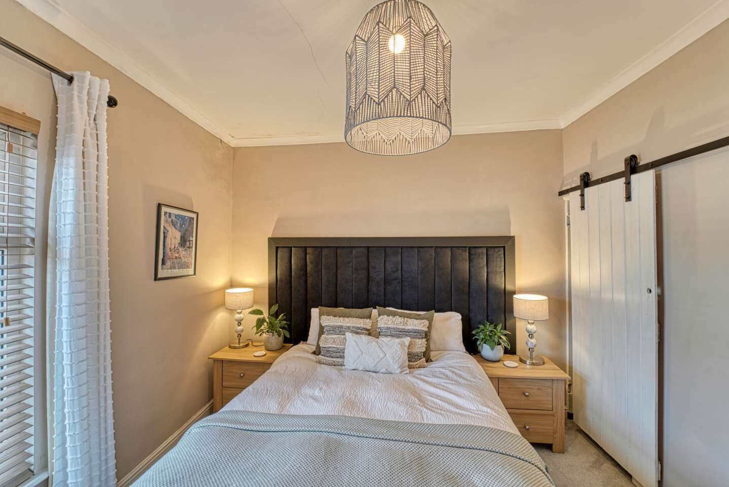 Quarto com uma cama grande, duas mesas de cabeceira, luminária pendente, janela com persianas e uma porta.