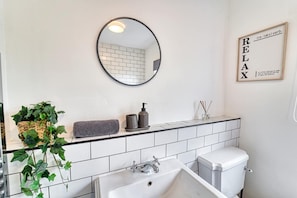 A modern bathroom with a white tiled backsplash, a round mirror, a sink, a toilet, and a potted plant. - Host Stay Birches Barn (Wolverhampton)