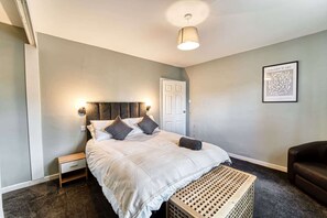Une chambre à coucher comprenant un lit, des tables de chevet, un banc en bois, un fauteuil en cuir foncé et une œuvre d’art encadrée accrochée au mur.
