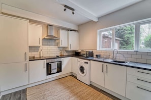 A compact kitchen with white cabinets, a built-in oven, a washing machine, and a window providing a view of greenery outside. - Host Stay Birches Barn (Wolverhampton)