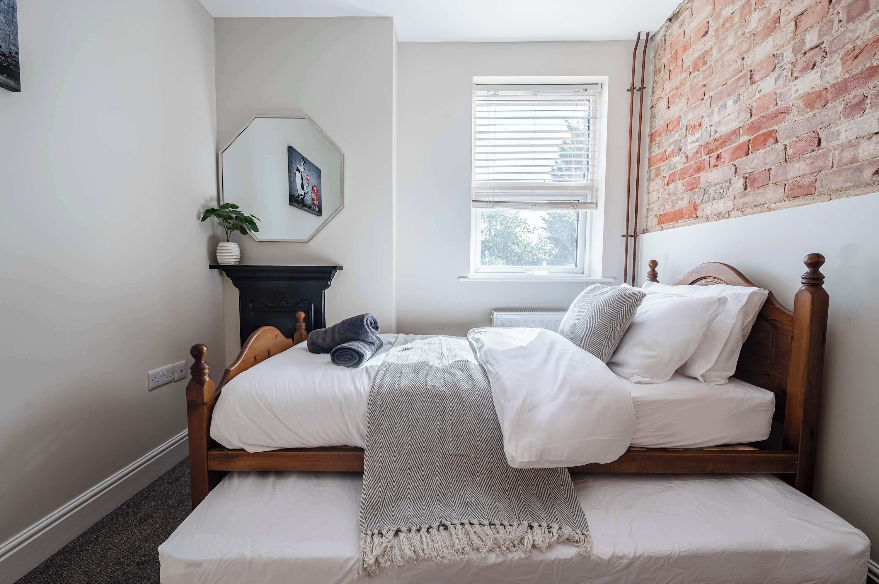 Une chambre avec un lit en bois, un mur blanc, un mur en briques, une fenêtre avec des stores et une petite plante posée sur une étagère.