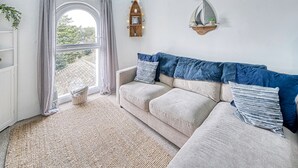 Un salon chaleureux avec un canapé beige, une grande fenêtre avec des rideaux, une étagère murale agrémentée d’objets décoratifs et un tapis à motifs.