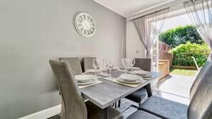 A dining area with a table set for four, a clock on the wall, and a view of a garden through a window. - Host Stay Walkers Lane House (Worcester)