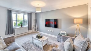 A modern living room with a flat-screen TV mounted on the wall, a white coffee table, grey sofas, a floor lamp, and a window with curtains. - Host Stay Walkers Lane House (Worcester)