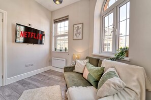 A cozy living room with a large flat-screen TV mounted on the wall, a comfortable sofa with multiple cushions, a small table with a lamp, and a window with a view of buildings outside. - Host Stay Taylor Bennett Apartment (Worcester)