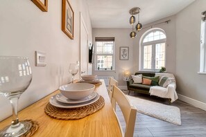 Un coin repas avec une table en bois, des assiettes et des bols blancs, un canapé vert et une fenêtre donnant sur des immeubles.