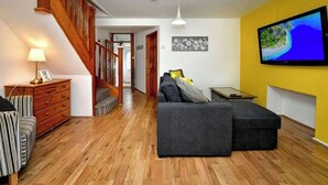 Uma sala de estar com escada de madeira, sofá cinza, televisão na parede e luminária em uma mesinha lateral.
