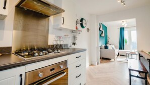 Eine moderne Küche mit Gasherd, weißen Schränken und einer Holz Arbeitsplatte. Es gibt einen Essbereich mit Tisch und Stühlen sowie ein Wohnzimmer mit Sofa und blauem Vorhang.