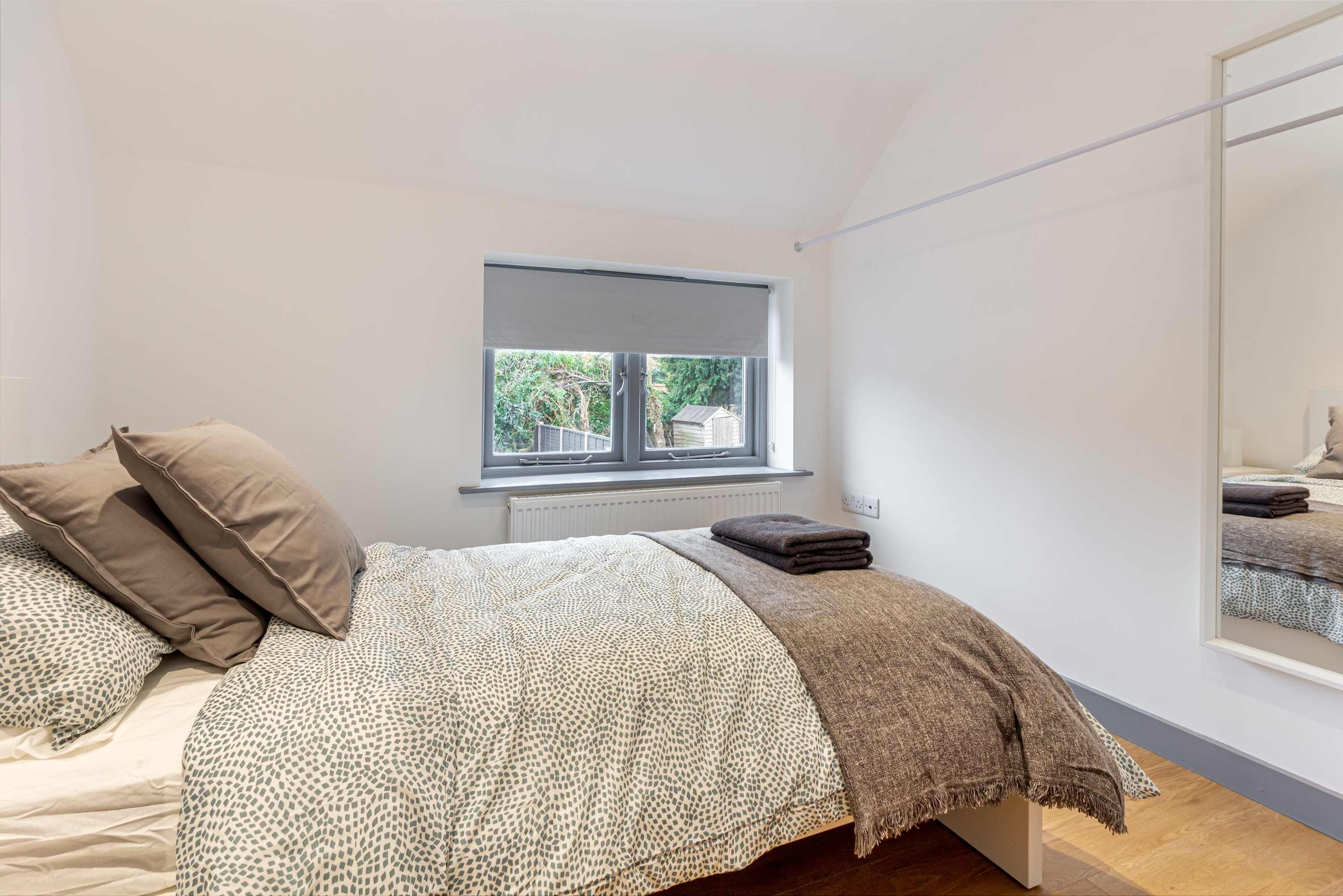 Een slaapkamer met een bed, kussens, een raam en een spiegel.