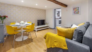 A modern living room with a grey sofa, yellow chairs, and a dining table with yellow chairs. There is a TV on a white stand, a small side table, and a painting on the wall. - Host Stay Bridge Street Residence (Leominster)