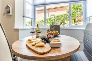 Una mesa redonda de madera con una tabla de quesos, copas de vino y una cesta de fruta.
