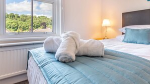 Ein ordentlich bezogenes Bett mit blauer Bettdecke und weißen Kissen, ein Fenster mit Blick auf einen Wald und eine Nachttischlampe.