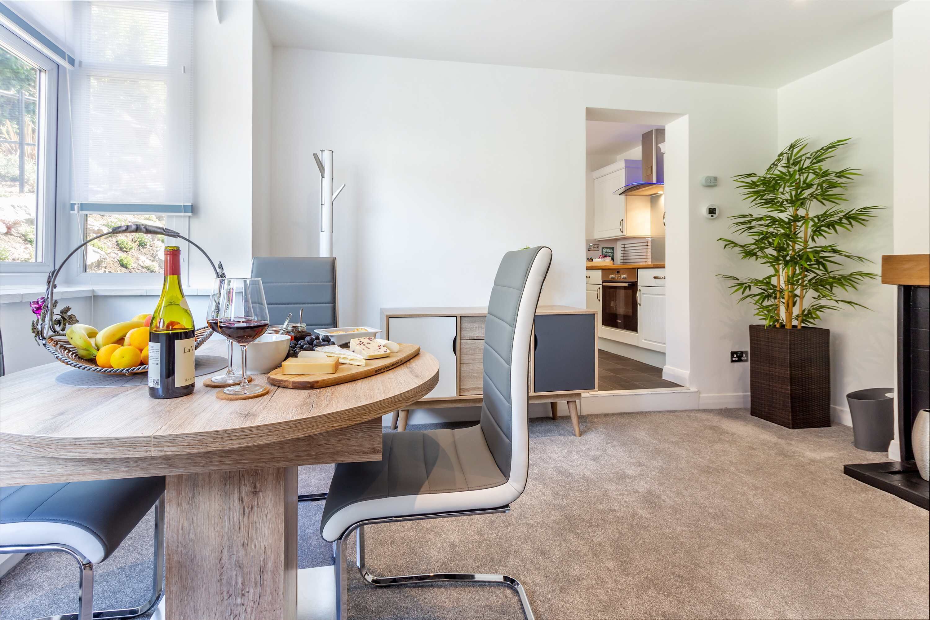 A modern dining area with a round table, chairs, a bottle of wine, and a fruit basket.