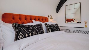 Cama bem arrumada com cabeceira laranja acolchoada, roupa de cama branca e almofadas estampadas. Uma mesa de cabeceira com abajur e espelho é visível.