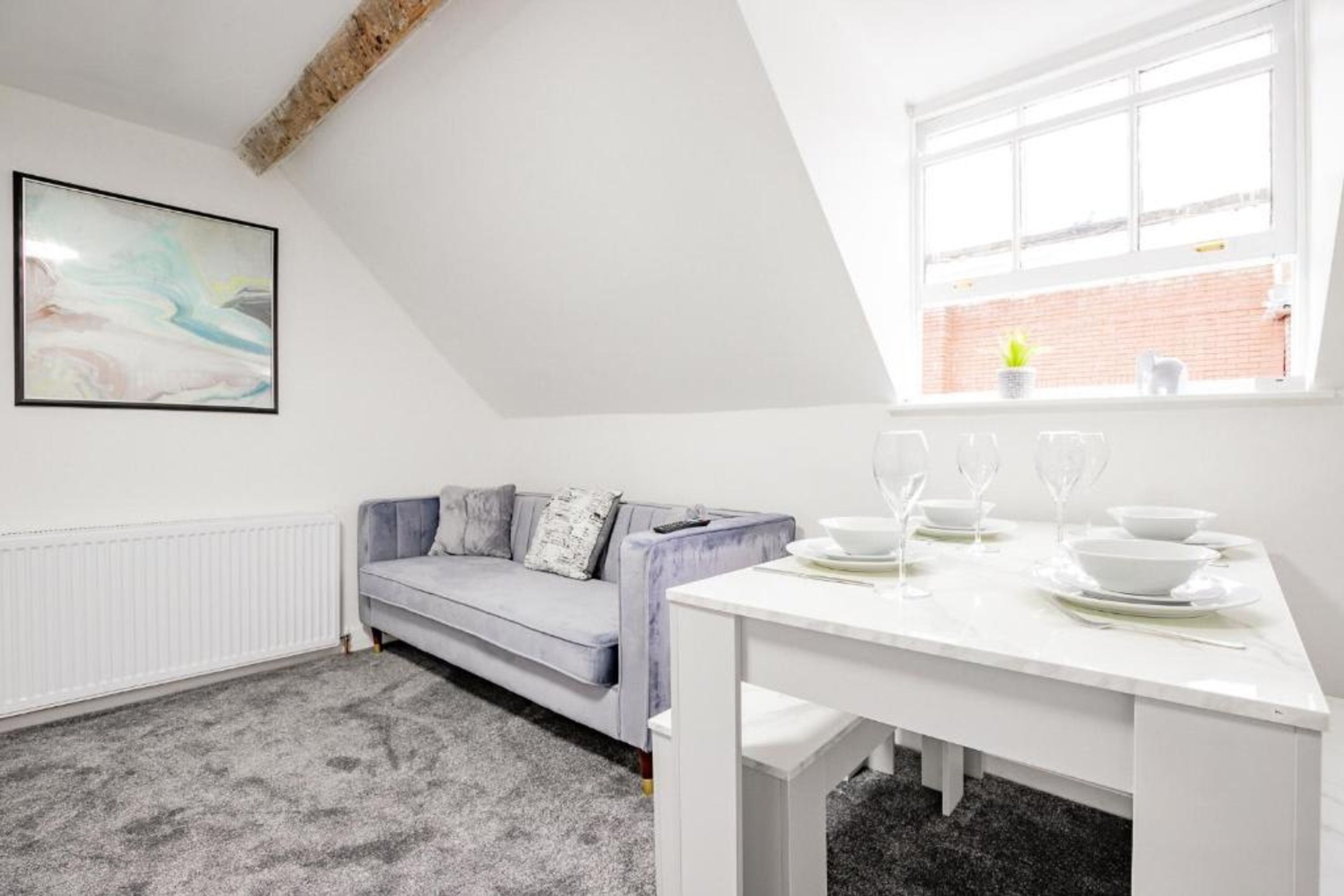 Une pièce moderne et bien éclairée, avec un canapé gris, une table à manger blanche mise avec des assiettes et des verres, un radiateur et une grande fenêtre donnant sur un mur de briques.