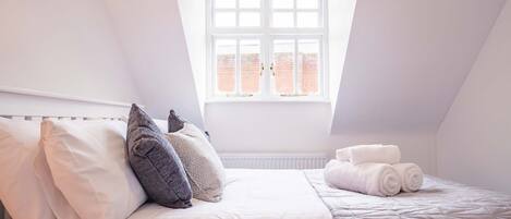 Quarto com teto inclinado, uma janela grande, cama com almofadas e toalhas dobradas.