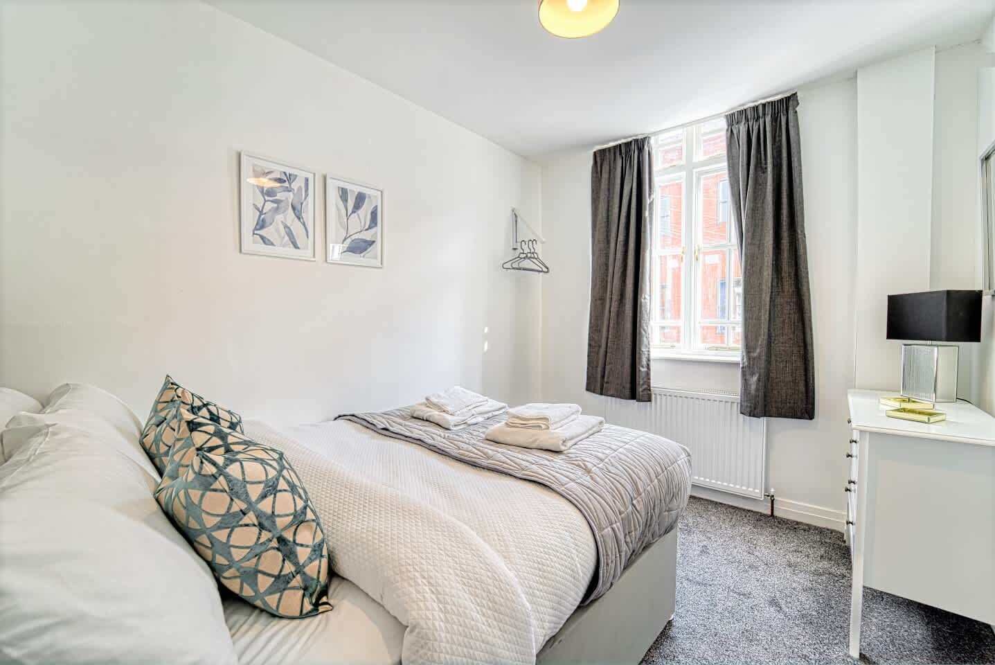 A bedroom with a bed, a bedside table with a lamp, a window with curtains, and two wall-mounted pictures.