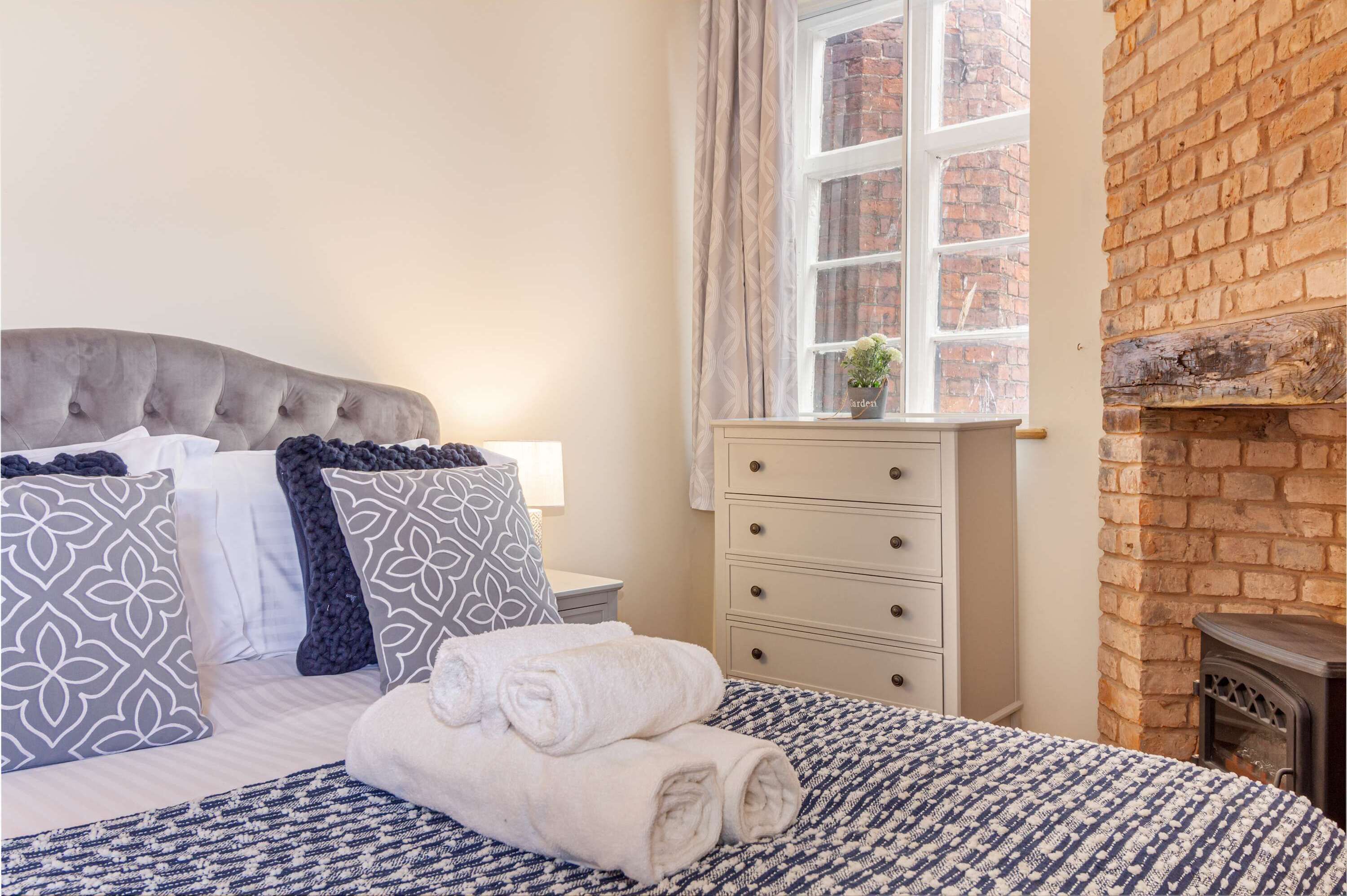 A bedroom with a tufted headboard, a bed with patterned bedding, a bedside table with a lamp, a dresser with drawers, and a brick wall.