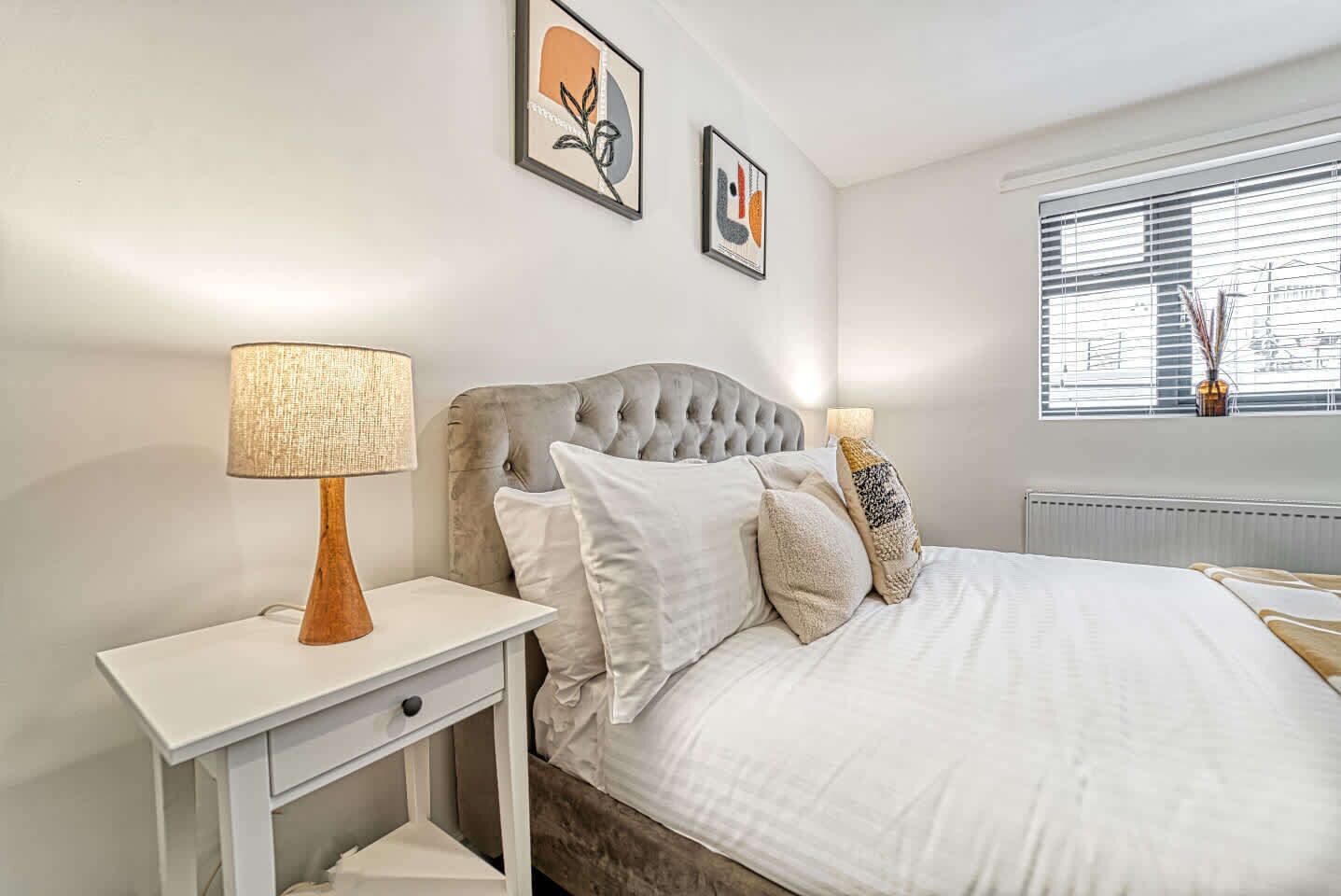 A bedroom with a tufted headboard, white bedding, a bedside table with a lamp, and two framed artworks on the wall.