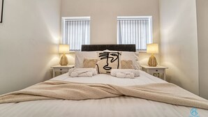 A neatly made bed with a large pillow featuring a face design, two bedside lamps, and a window with blinds. - Host Stay The Bell (Stourbridge)