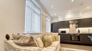 A modern living room with a beige sofa, a kitchen with dark cabinets, and a large window with blinds. - Host Stay The Bell (Stourbridge)