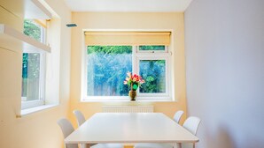 Een eethoek met een witte tafel en stoelen, een raam met uitzicht op groen, en een vaas met bloemen op de vensterbank.