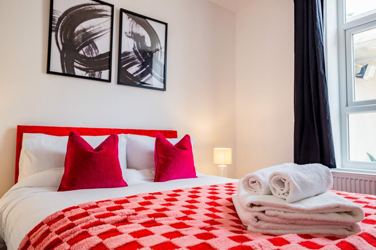 A hotel room with a bed, red and white checkered blanket, two red pillows, and two abstract paintings on the wall.
