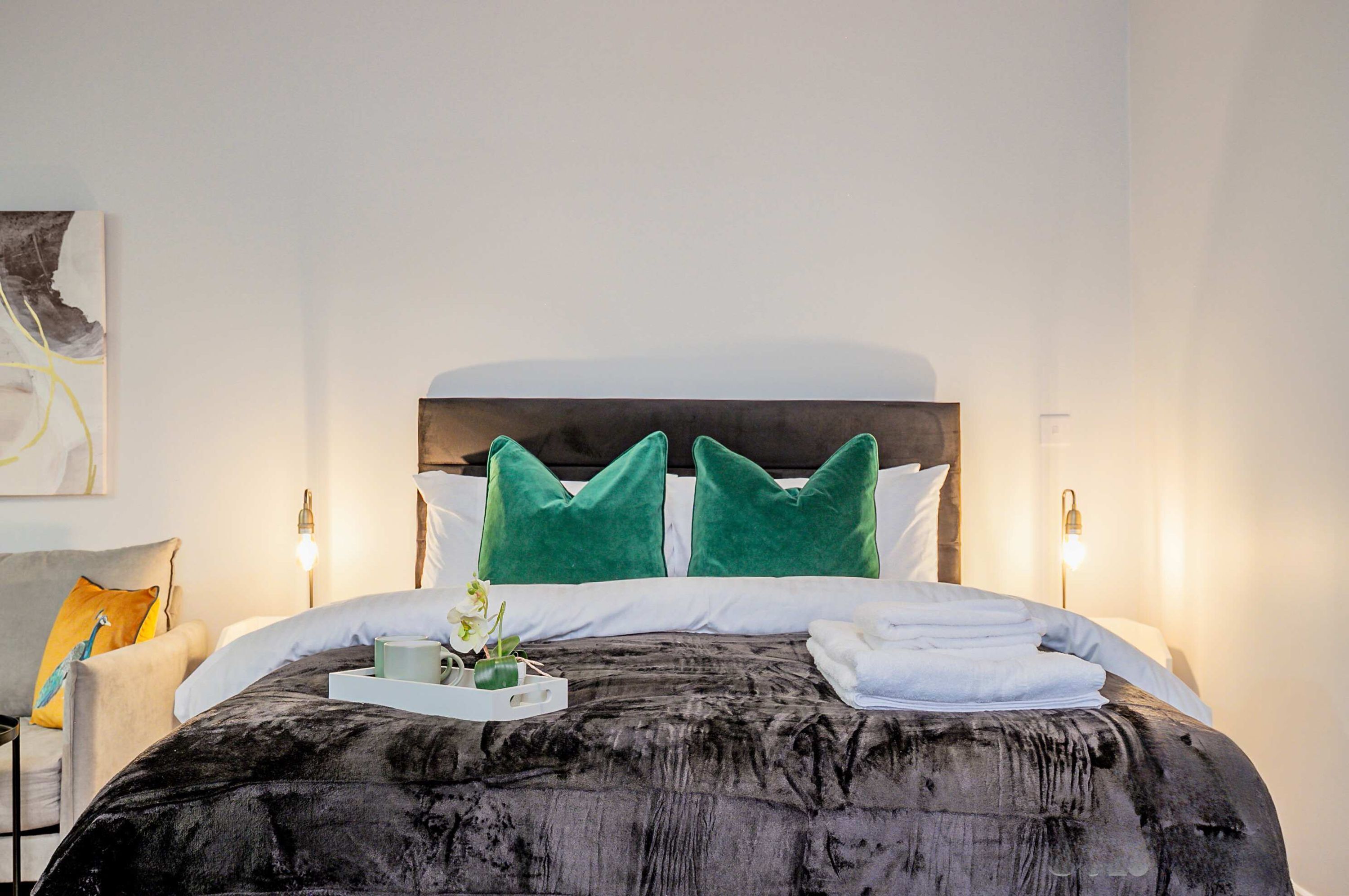 A neatly made bed with a dark, textured bedspread, two green pillows, and a white headboard. There is a bedside table with a lamp and a tray with a cup and flowers.