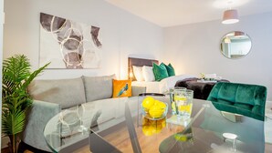 Ein modernes Wohnzimmer mit einem Glastisch, einer Couch, einem Esstisch mit einer Zitronenschale und Gläsern sowie einem grünen Sessel.