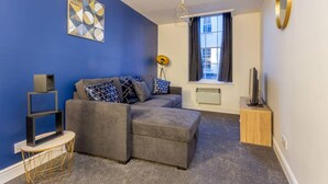 Ein modernes Wohnzimmer mit einem grauen Ecksofa, einem kleinen runden Beistelltisch, einer Stehlampe und einem großen Fenster mit Vorhängen.