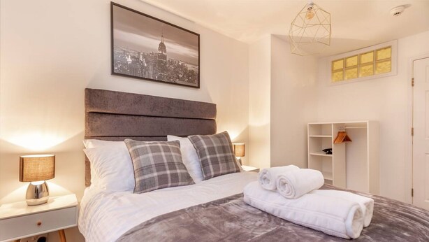 Ein ordentlich bezogenes Bett mit einer grauen Decke, zwei Kissen und einem aufgerollten weißen Handtuch. Ein Nachttisch mit Lampe, ein an der Wand befestigtes Regal mit Kleidung und ein gerahmtes Stadtbild über dem Bett.