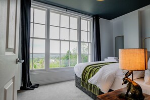 Um quarto com uma janela grande, uma cama com roupa de cama branca, um cobertor verde, uma mesa de cabeceira de madeira com uma luminária e uma estátua de uma raposa.